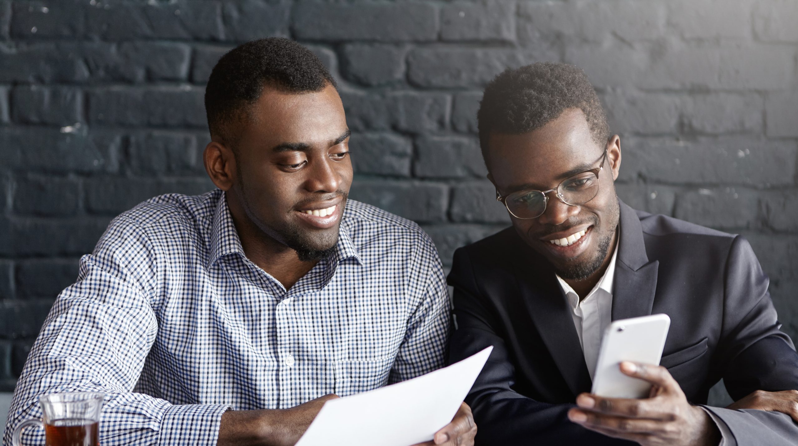 People, cooperation and teamwork concept. Two happy confident dark-skinned colleagues surfing internet on mobile phone while having break during brainstorming session, sharing business ideas